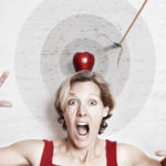 A woman with a surprised and scared expression on her face make eye contact with the camera.  An apple sits on top of her head and an arrow pierces a target behind her.